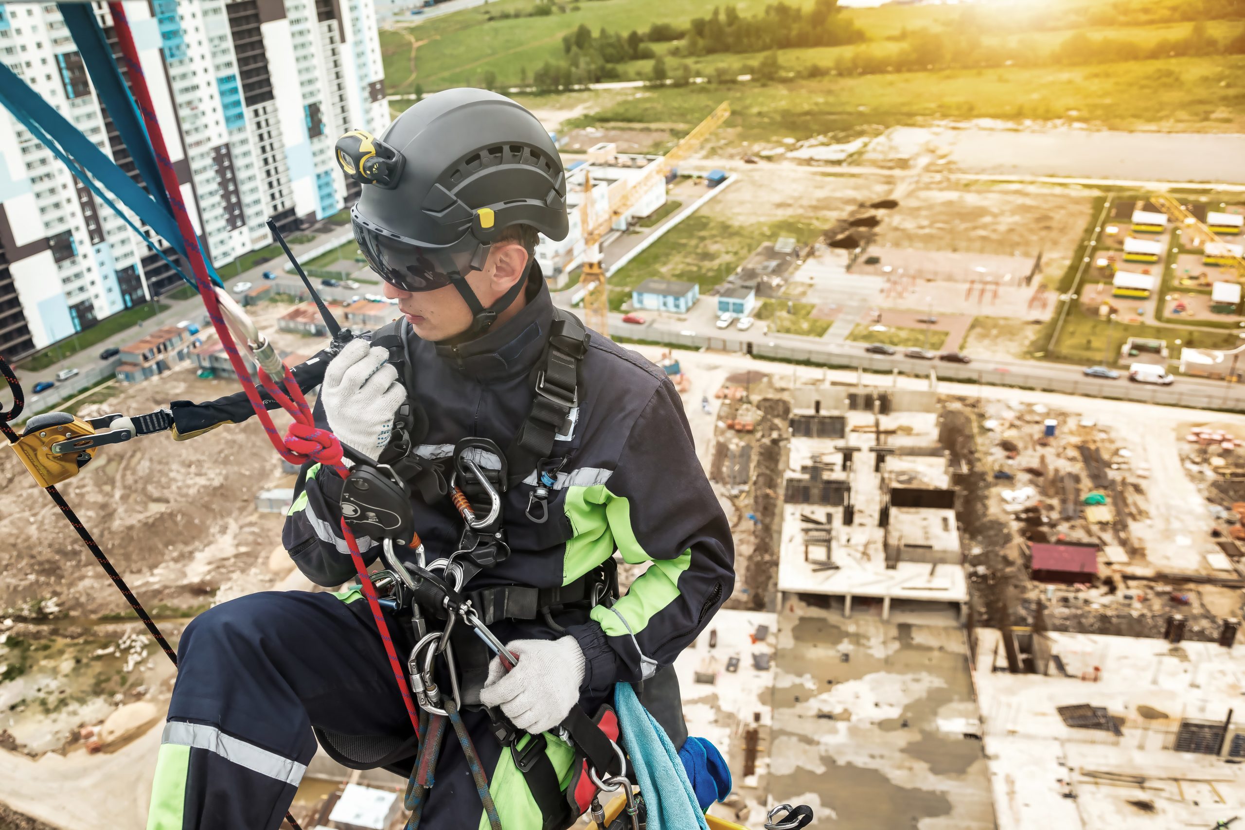 Industrial mountaineering worker in uniform talking walkie-talkie hangs on wall skyscraper at buildings backdrop. Rope access laborer during high-rise work. Concept of industry urban works. Copy space